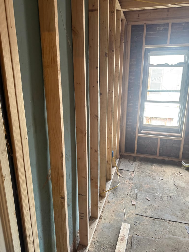 Interior construction site with wooden wall studs and framing, and window opening with light streaming in