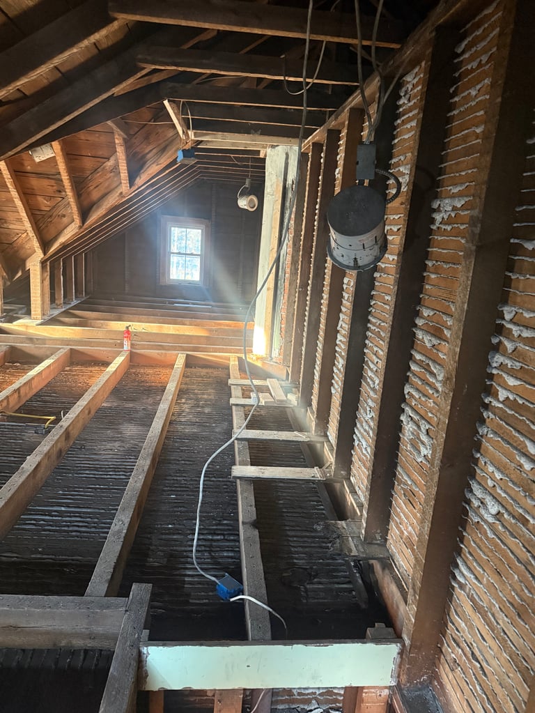Interior of an old attic with exposed wooden beams, brick walls, and a small window letting in light