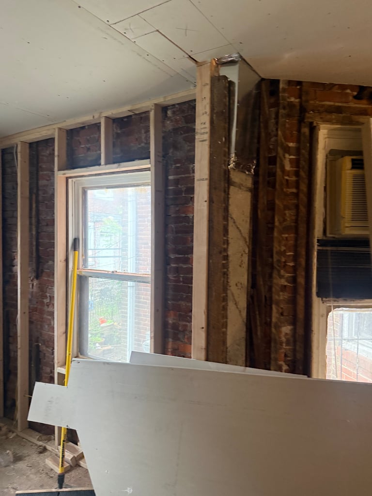 Interior renovation in progress showing exposed brick walls, wooden framing studs, window opening, and drywall installation