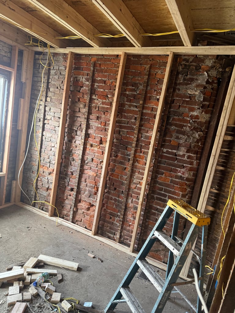 Construction interior showing exposed brick walls framed with wooden studs, exposed ceiling beams, and a ladder on the floor