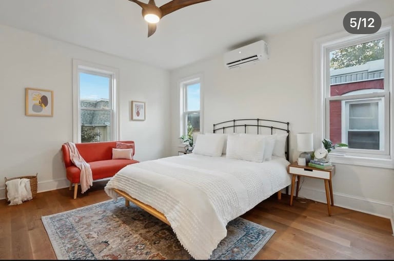 Bright modern bedroom with hardwood floors, queen bed with white bedding, red accent chair, and large windows overlooking trees