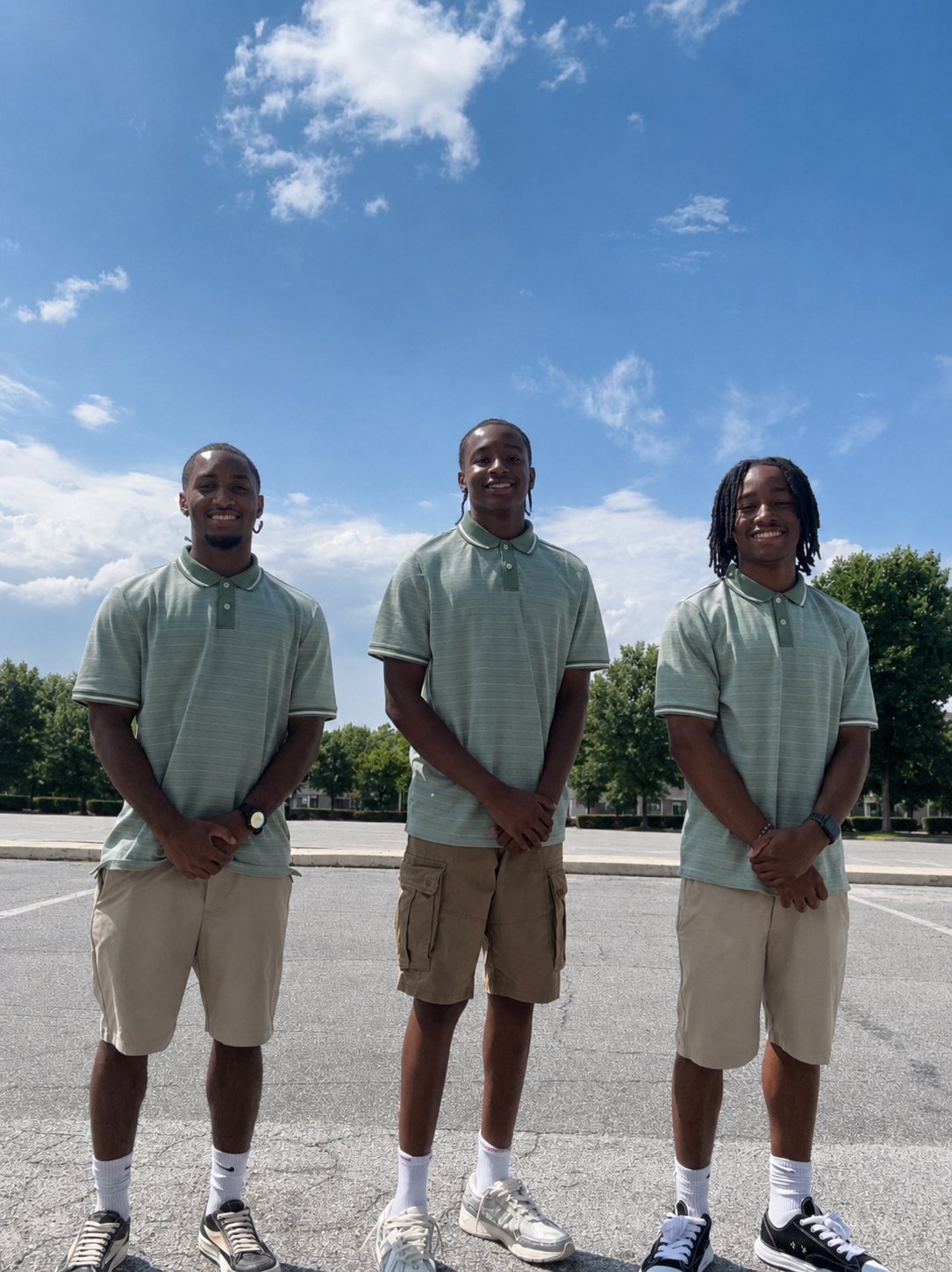 Three men in matching light green polo shirts and khaki shorts standing together on a paved area under a blue sky with white clouds
