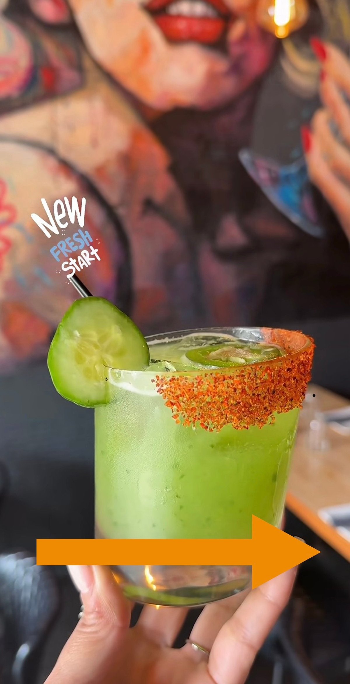Bright green cocktail with red spiced rim and cucumber slice, garnished with New Fresh Start text and orange arrow, held in hand