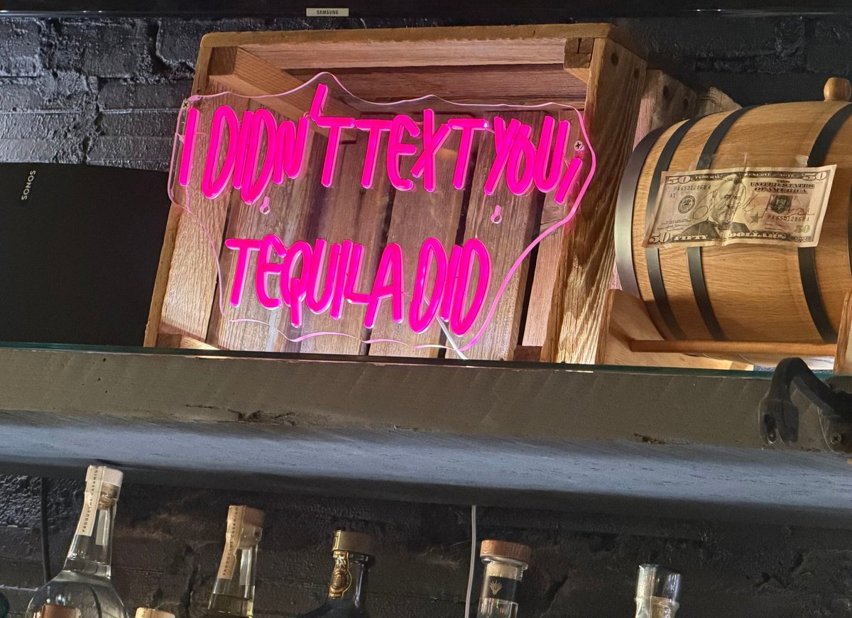 Bright pink neon sign reading I Didn't Text You, Tequila Did displayed on a wooden shelf with vintage decor, wooden barrel, and tequila bottles below