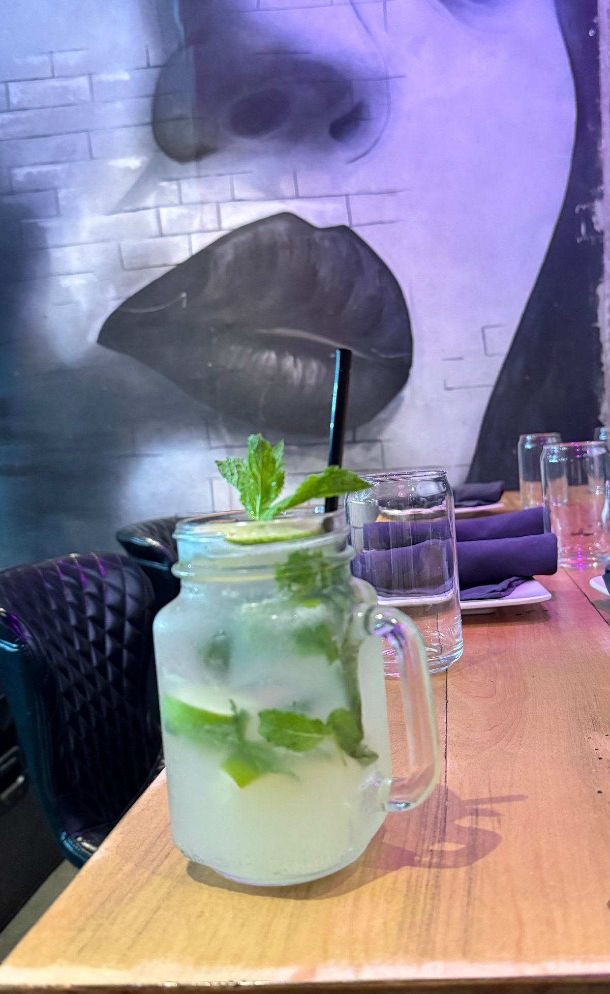 Mojito cocktail with fresh mint and lime in a glass pitcher on a wooden table, with purple lip art mural in background