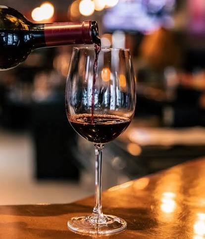 Red wine being poured from a bottle into a wine glass on a wooden bar with warm bokeh lights in the background