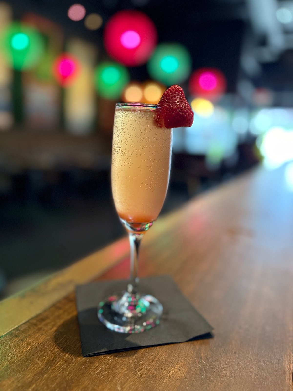 Champagne cocktail with fresh raspberry garnish on black coaster, colorful blurred lights in background at upscale bar