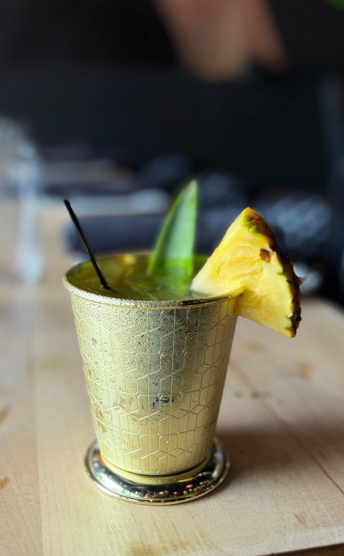 Yellow cocktail in textured glass with pineapple slice, black straw, and fresh mint garnish on wooden surface