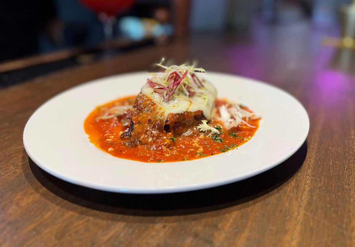 Gourmet plated Maduro Lasagna — sweet plantain lasagna with braised meat in orange pomodoro sauce, topped with shredded vegetables, served on a white plate