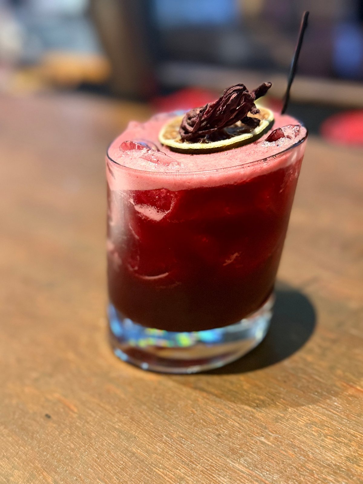 A vibrant red cocktail with ice, topped with a lime slice and chocolate garnish, served in a glass on a wooden surface