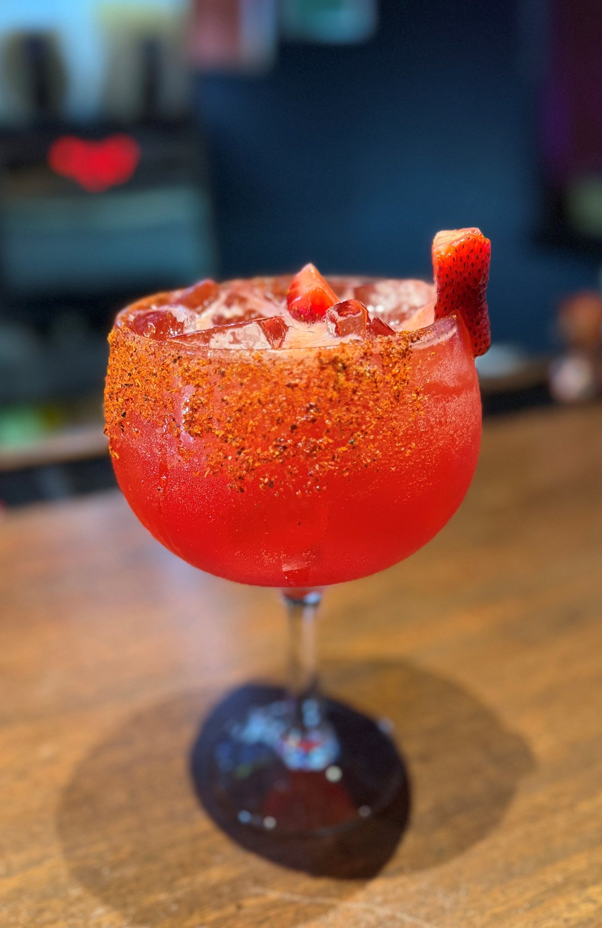 La Bichota jumbo margarita — red cocktail with chili-sugared rim, ice, and a strawberry garnish, served in a coupe glass on a wooden bar