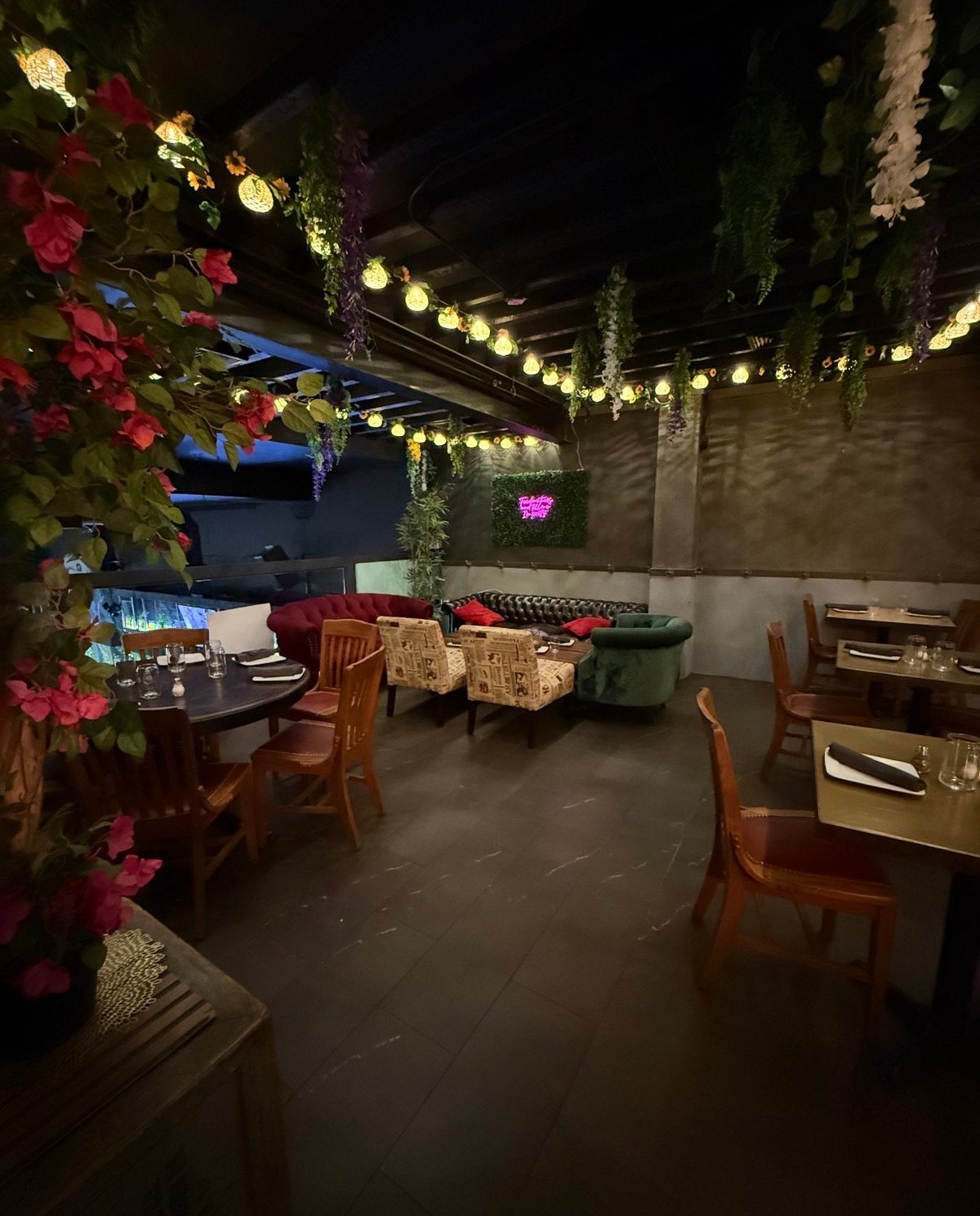 Nighttime rooftop terrace with string lights, pink flowers, wooden seating areas, and cushions overlooking the city