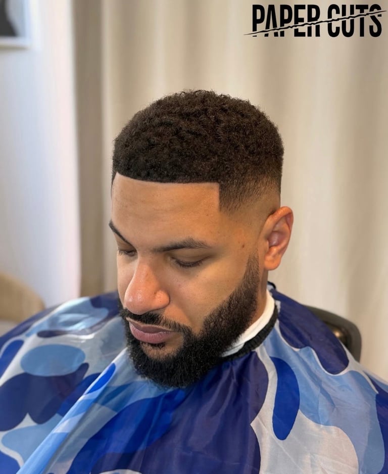 Man with short textured haircut and full beard wearing blue patterned cape, looking downward in professional barber shop setting