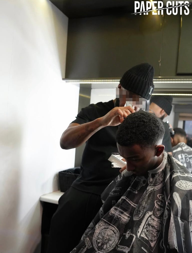 Barber giving a haircut to a client in a salon, Paper Cuts logo visible in top right corner
