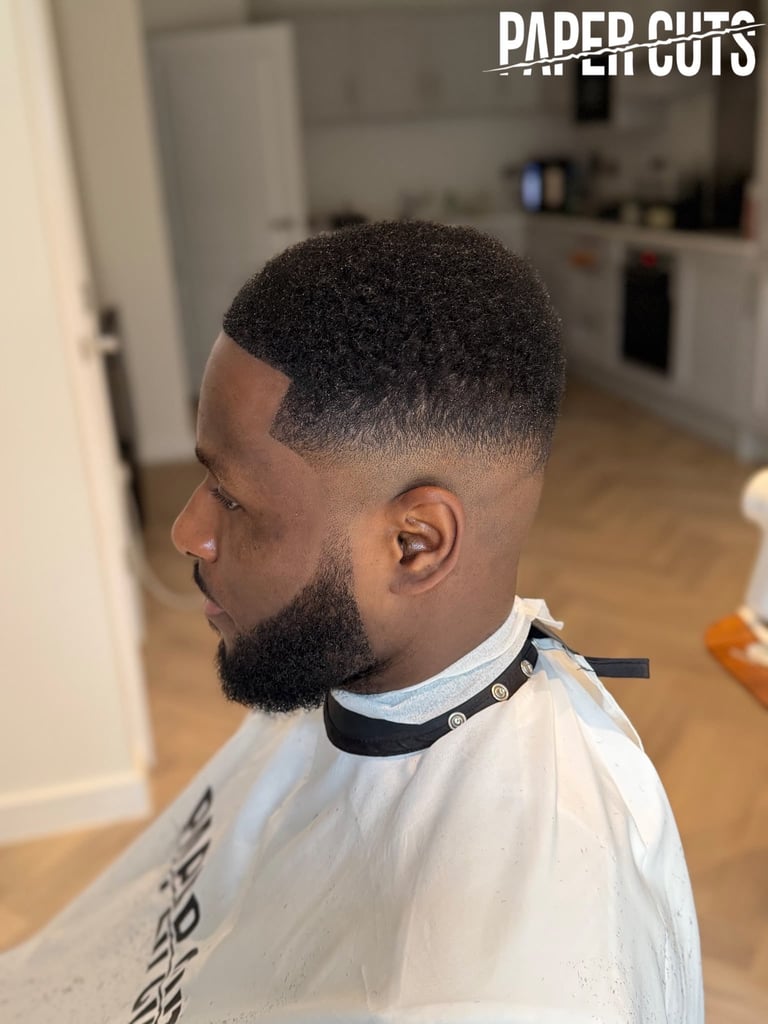 Man with faded fade haircut and full beard shown in profile view wearing barber cape in salon setting