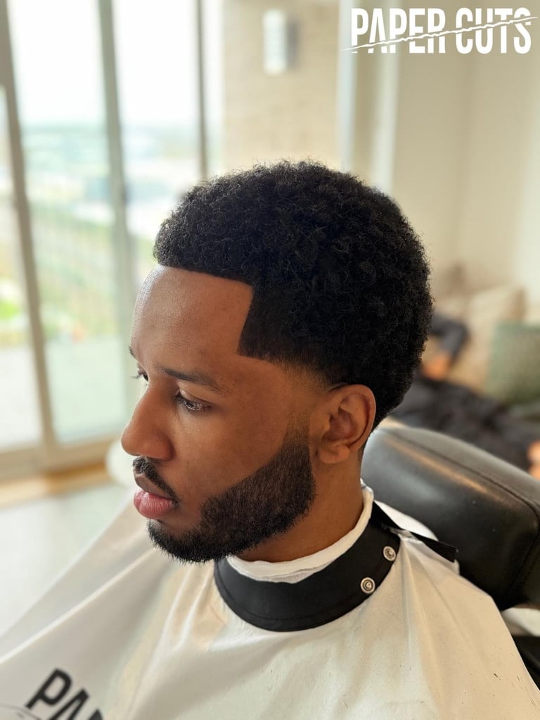 Man with fade haircut and beard in barber chair, viewed in profile with salon interior and window visible in background