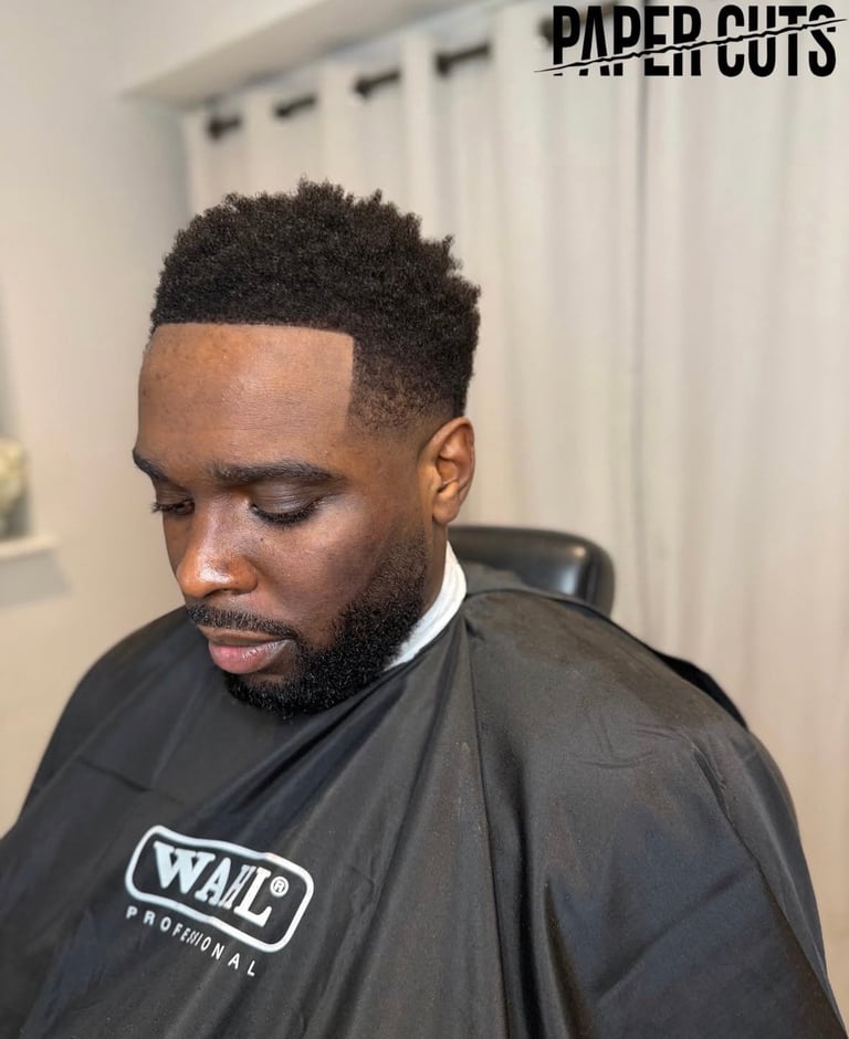 Man with fade haircut and beard wearing black barber cape labeled Wahl Professional, shown in profile inside a salon