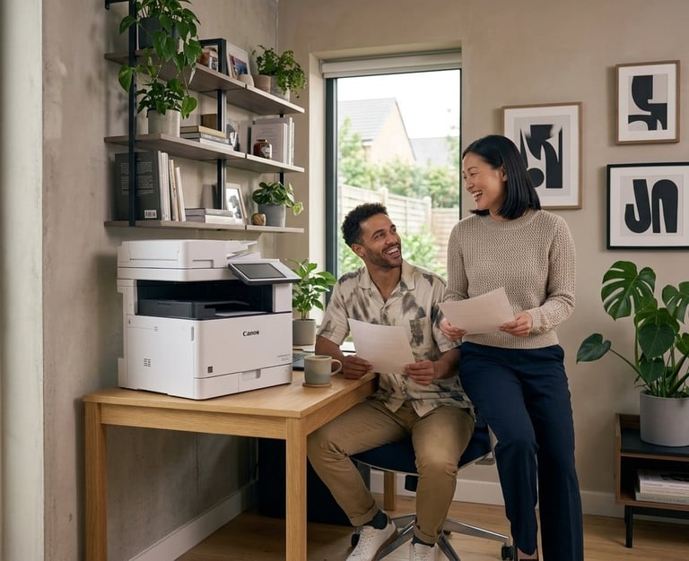 Two people reviewing documents in a home office with a printer on a wooden desk, shelving, plants, and abstract wall art.