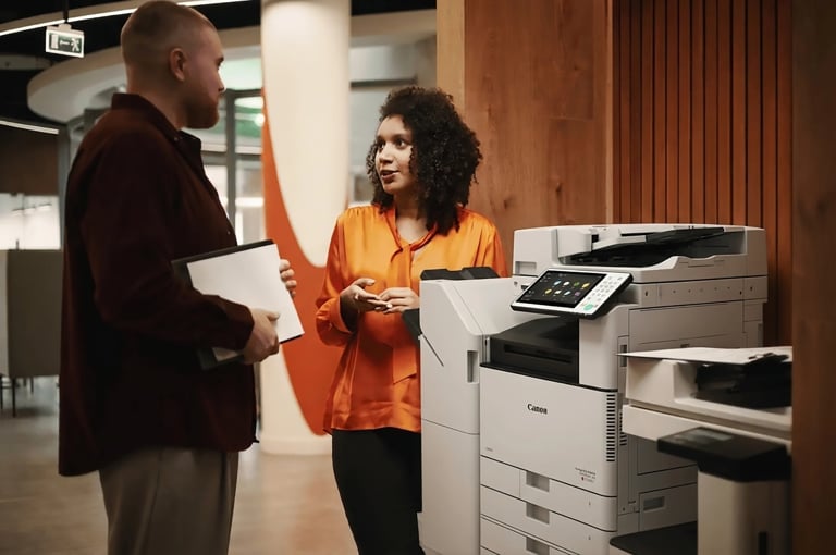 Woman in orange shirt speaking with man at office multifunction printer in modern workplace
