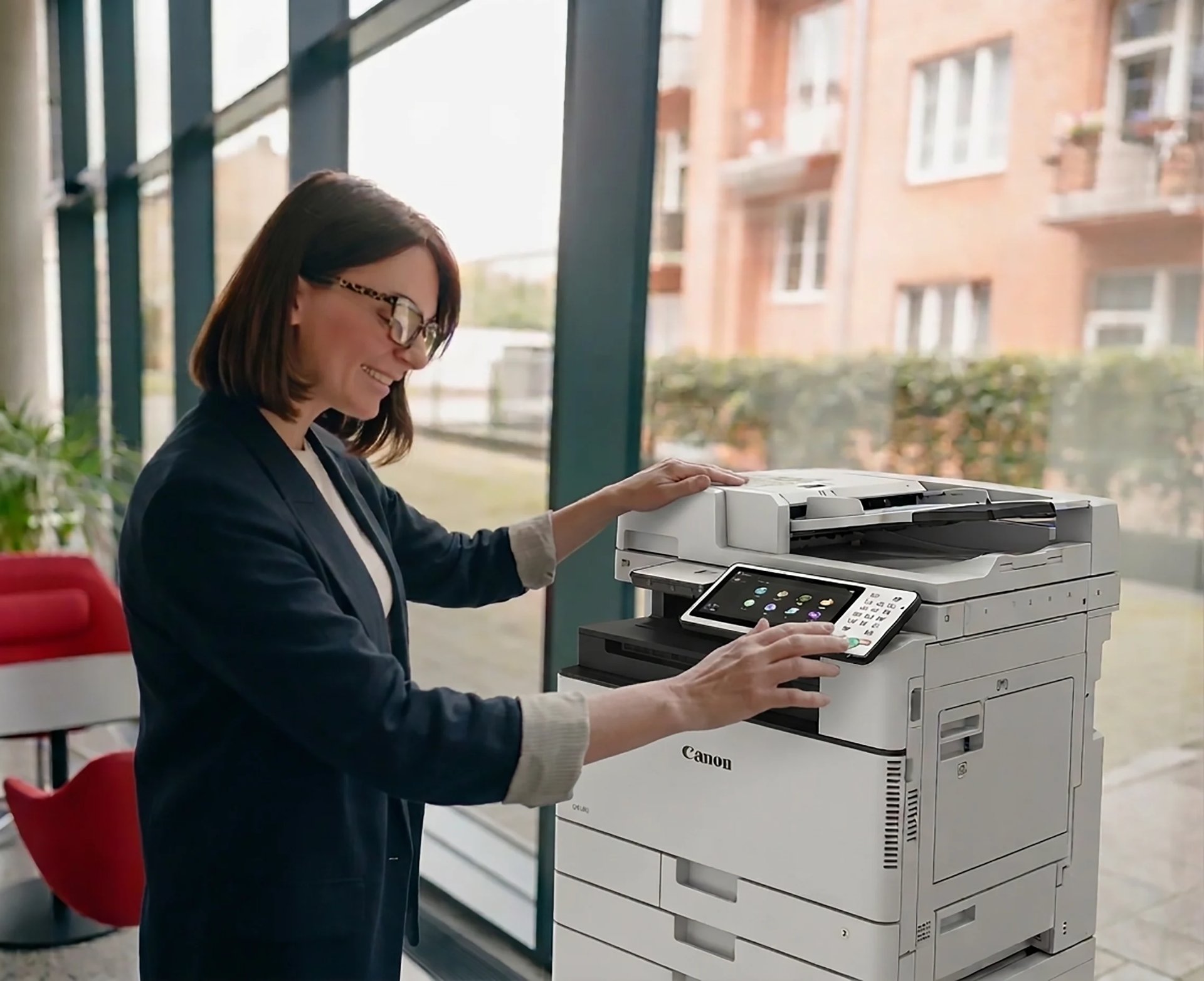 Woman in business attire using a Canon multifunction printer in a modern office with windows and city buildings visible in background.