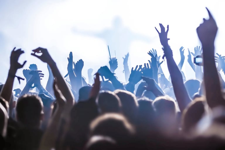 Silhouetted crowd at a concert or music festival with raised hands and arms celebrating, shot from ground level against a bright blue sky