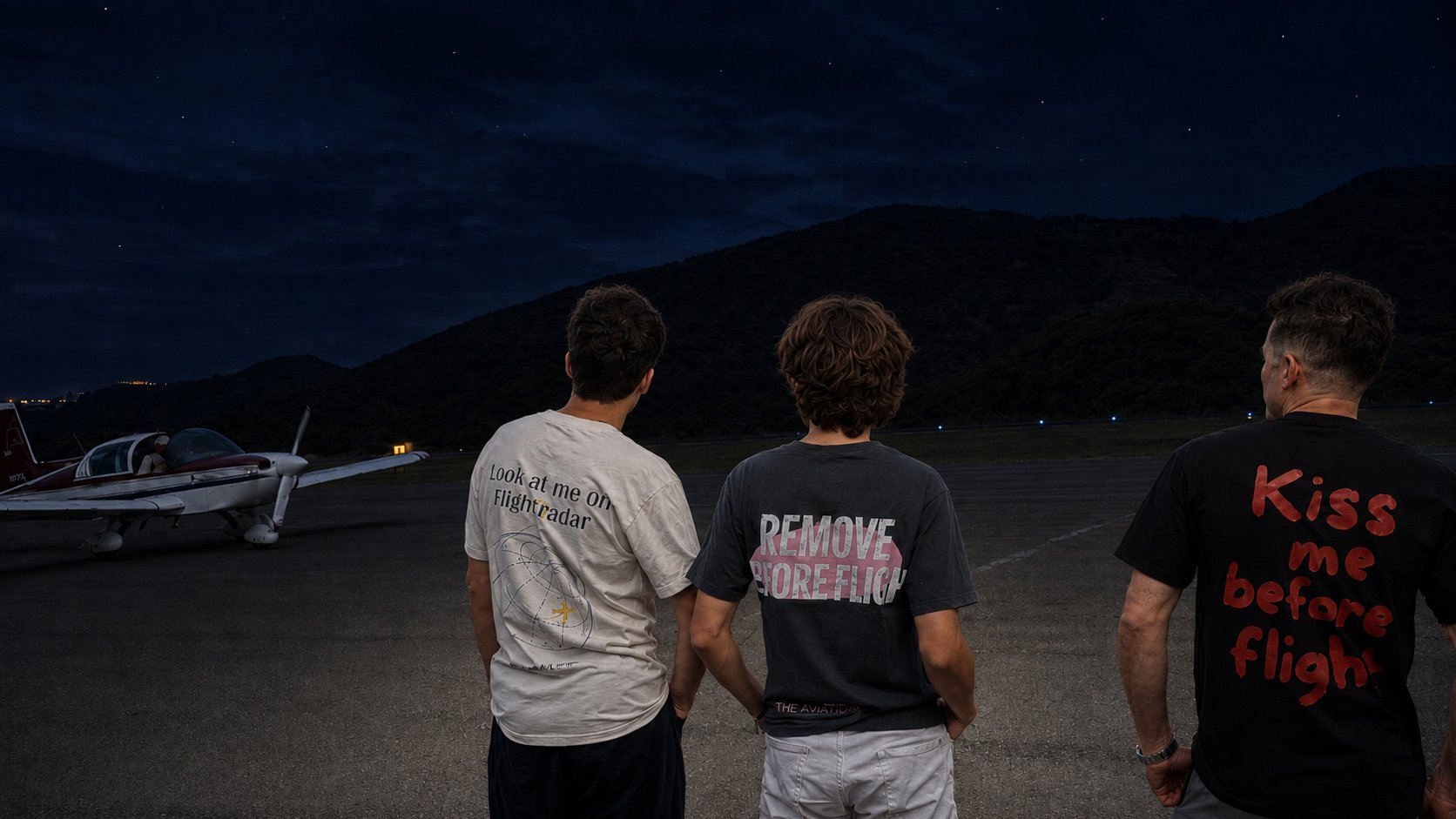 Three men standing on a runway at dusk facing an airplane in the distance with mountains in the background