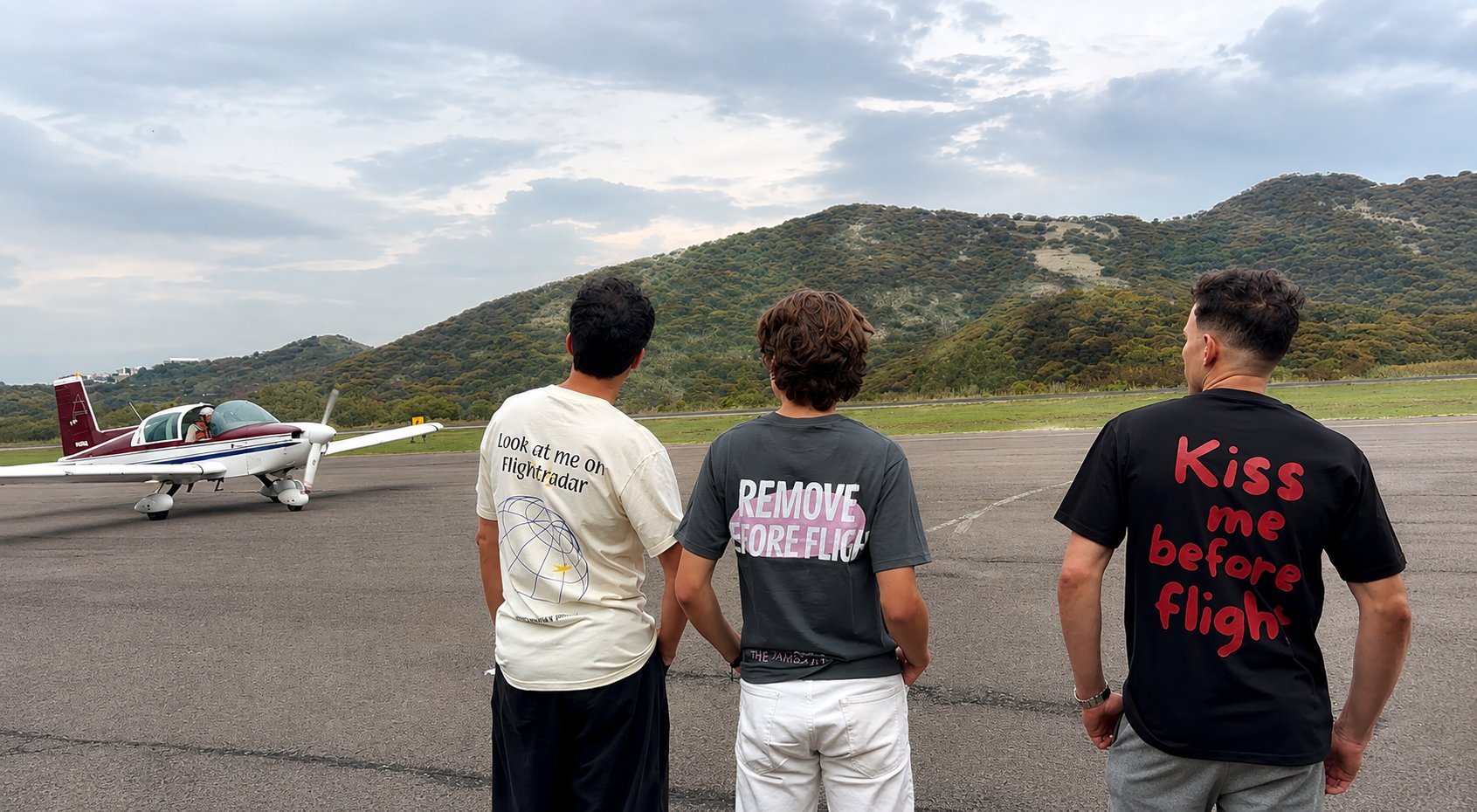 Three men from behind standing on an airstrip, watching a small airplane on the runway with mountains visible in the background.