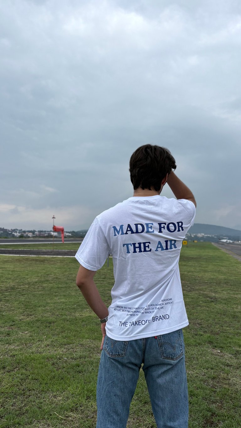 Person wearing a white Made for the Air t-shirt standing in an open field, shielding eyes while looking toward an airport runway