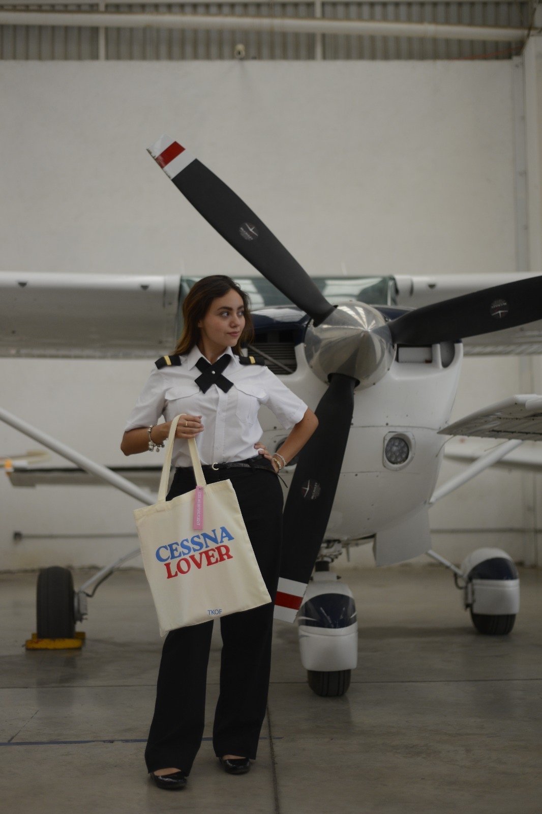 Woman in pilot uniform holding a canvas tote bag standing in front of a parked aircraft in a hangar