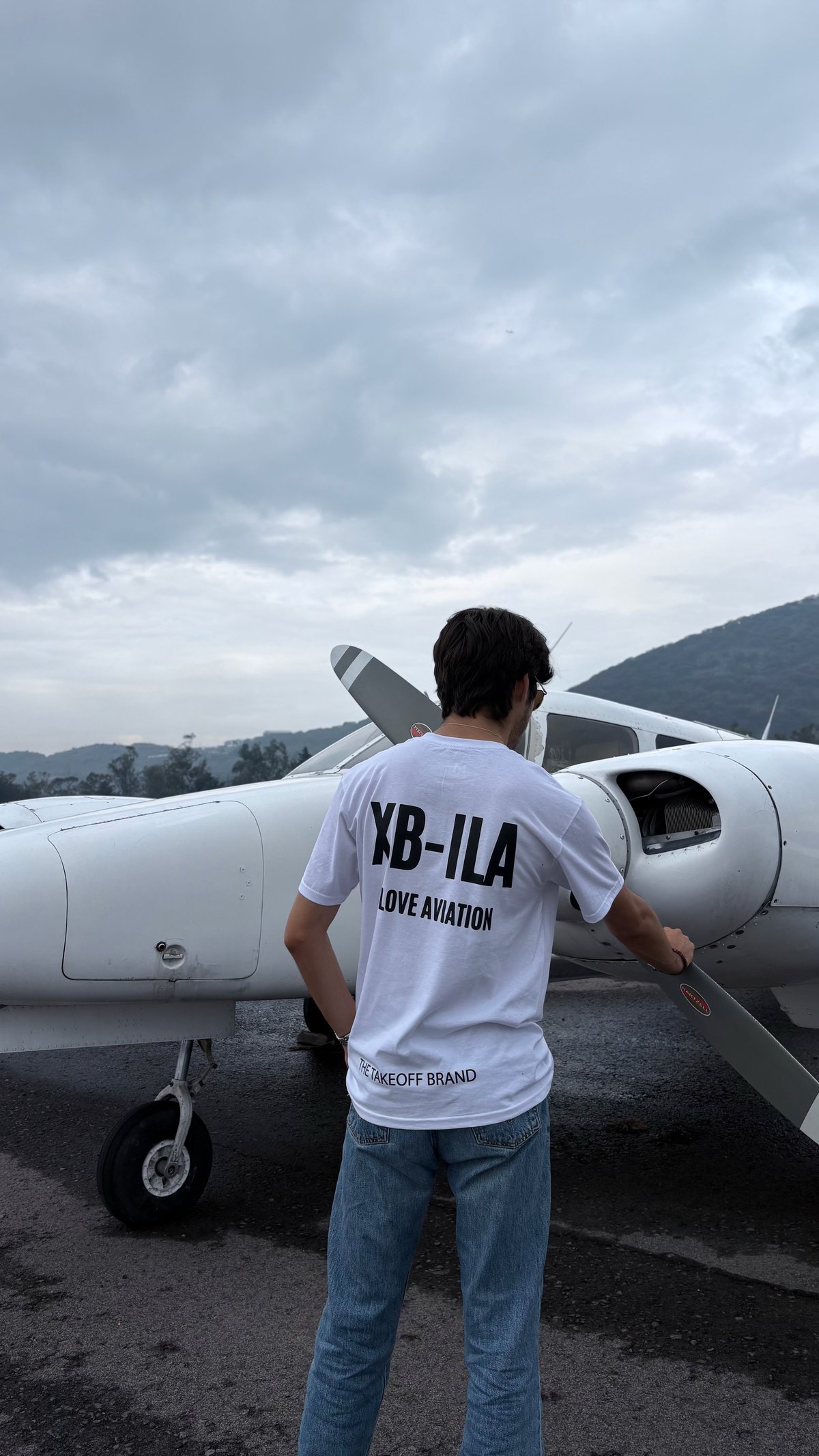 Person in white XB-ILA t-shirt standing in front of small aircraft with mountains in background