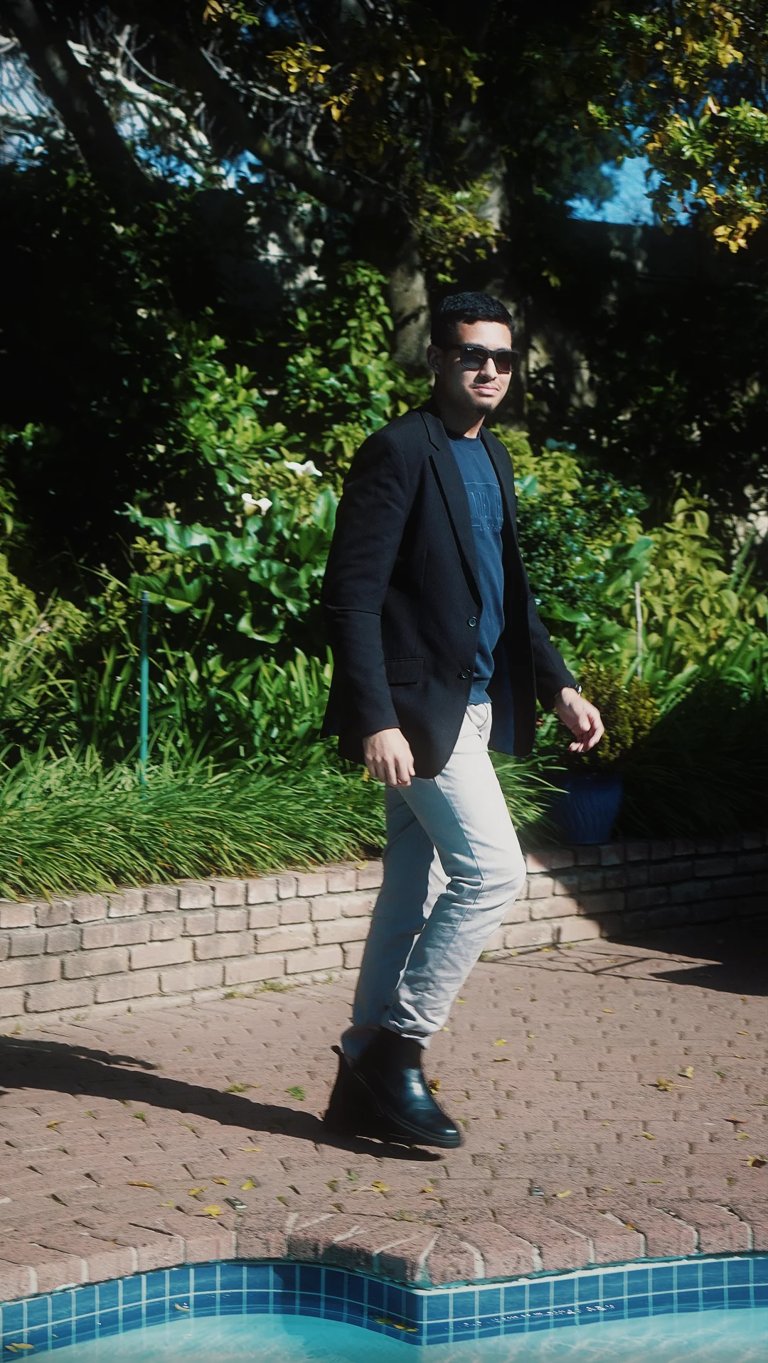 Man in dark blazer and light jeans standing on brick patio by pool, surrounded by green trees and blue sky