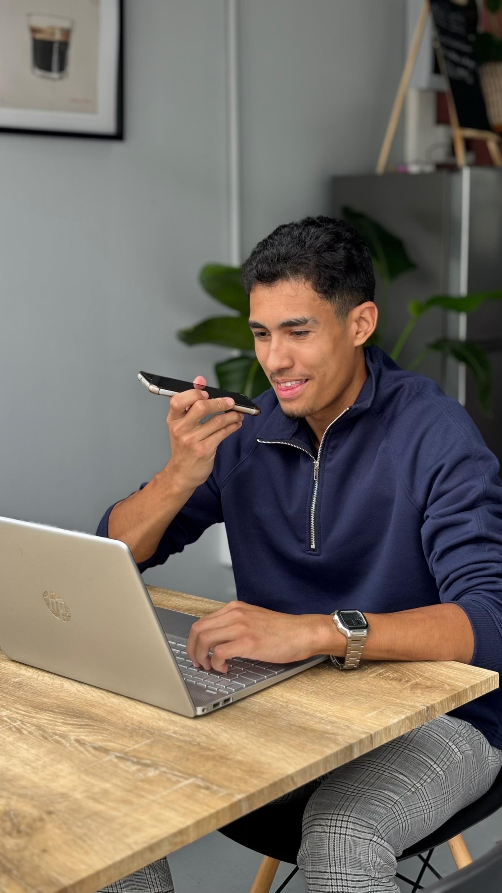 Man using voice command on smartphone while working at laptop at wooden desk in modern office space