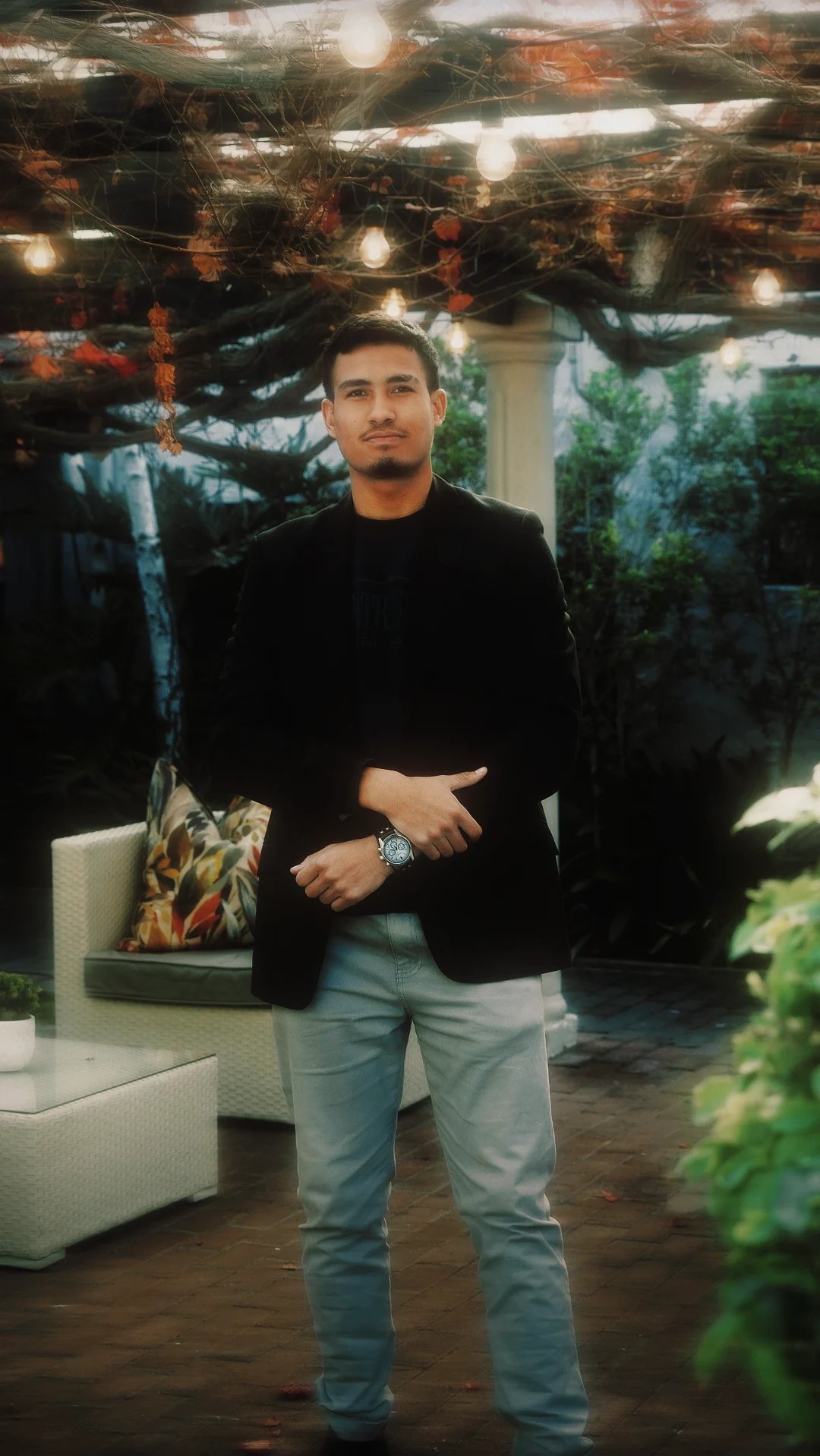 Man in black blazer and jeans standing in a decorated outdoor patio with string lights and greenery