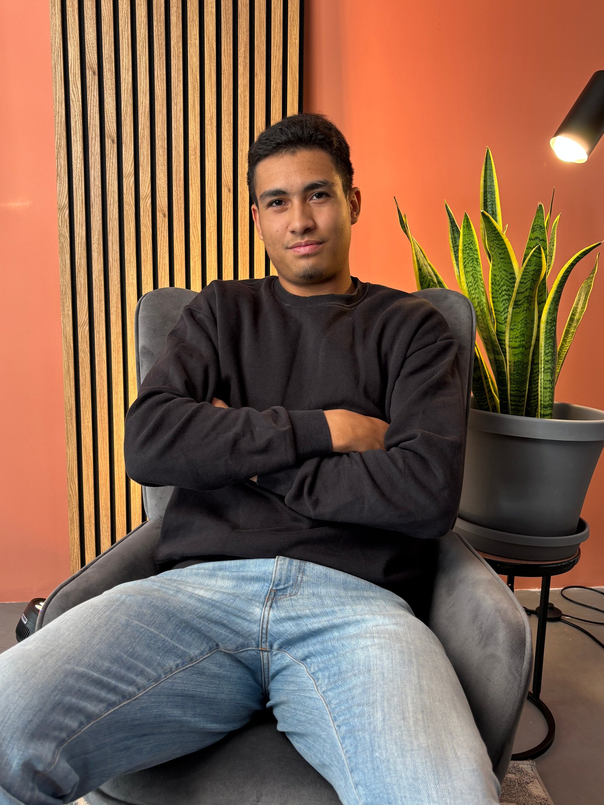 Man sitting in gray chair with arms crossed, wearing black sweatshirt and jeans, in front of orange wall with vertical slats and potted snake plant