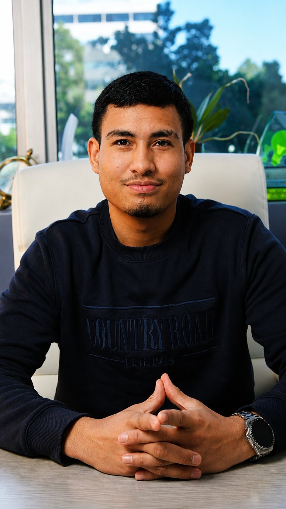 Man in black sweatshirt sitting at desk with hands clasped, smiling at camera by window with outdoor view