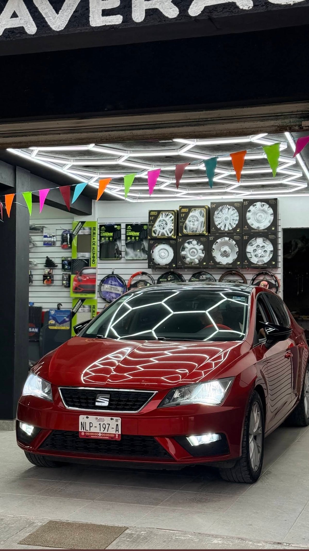 Auto rojo exhibido en Suki Motors Garage, con luces LED hexagonales en el techo, banderines de colores y exhibidores con tapones y rines en el fondo