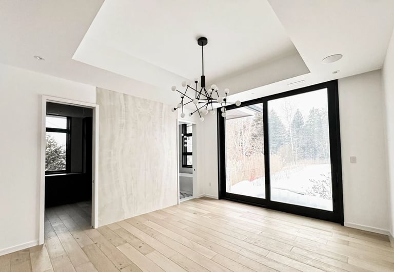 Modern minimalist interior with light wood flooring, white walls, black-framed glass doors and windows overlooking snow-covered landscape