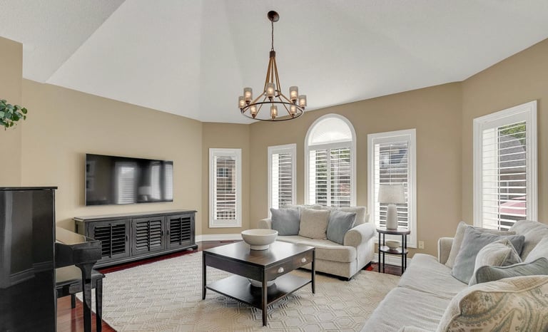 Bright modern living room with beige walls, white shuttered windows, black TV console, gray furniture, and decorative chandelier