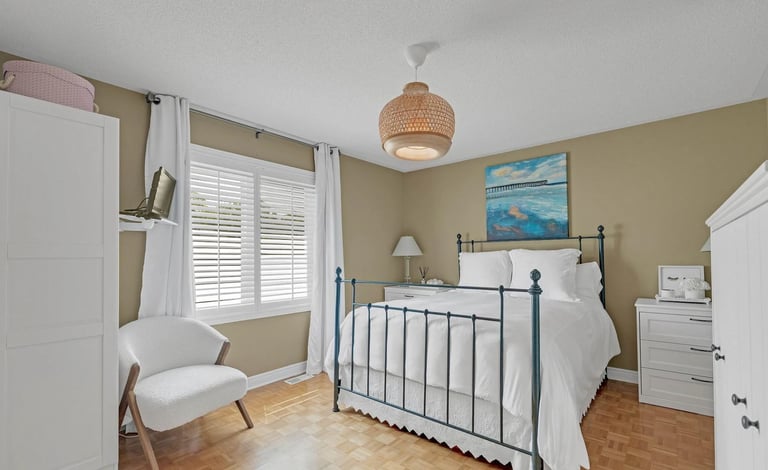 Bright bedroom with black metal bed frame, white bedding, beige walls, hardwood floor, white shuttered window, rattan pendant light, and white furniture