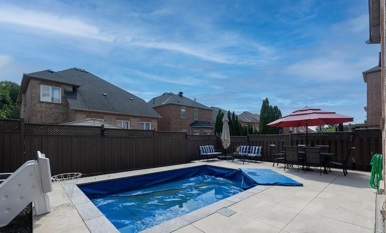 Backyard swimming pool with blue water surrounded by concrete patio, black fence, lounge chairs, and residential homes under clear blue sky
