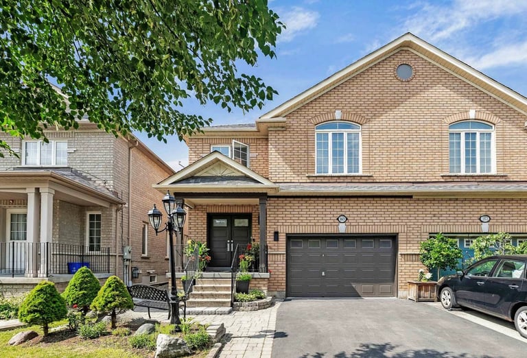 Two-story brick suburban home with dark garage doors, stone driveway, and lush green landscaping on a sunny day