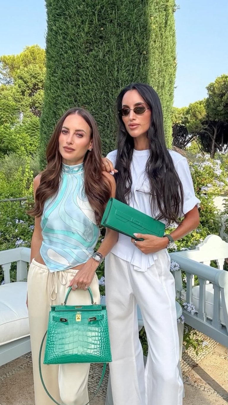 Two women posing outdoors with green accessories. Left woman wears light blue top and beige pants with green handbag. Right woman wears white outfit and sunglasses with green clutch.