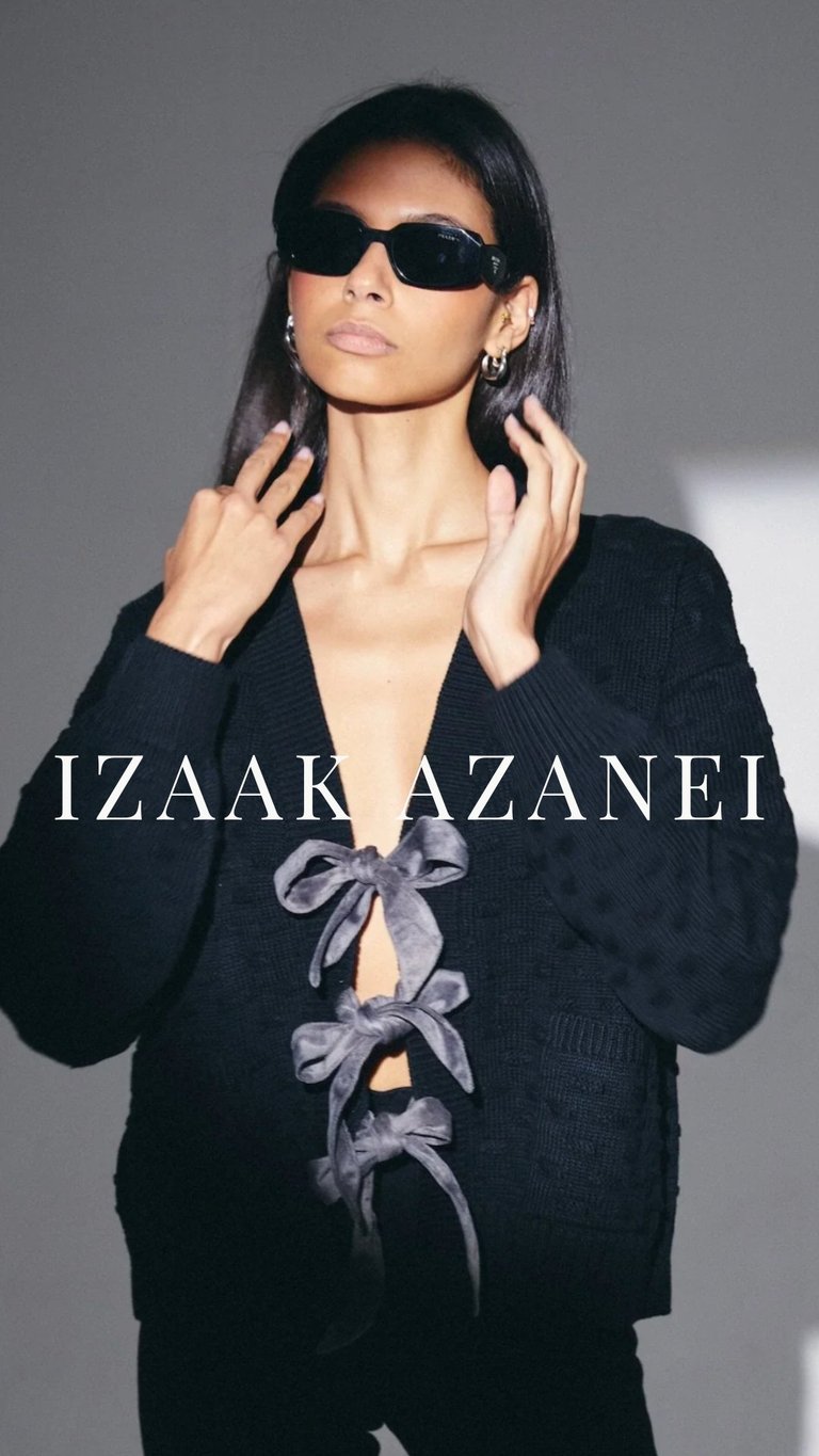 Woman wearing black sunglasses and black cardigan with ribbon details, hands near face, against gray background with IZAAK AZANEI text overlay