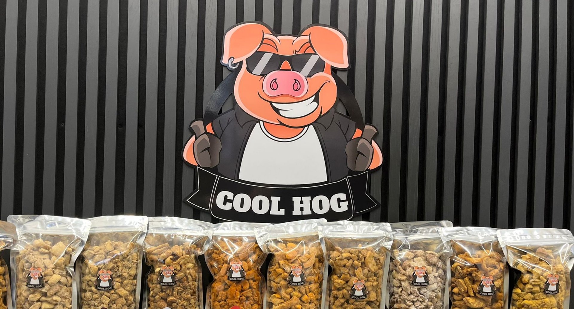 Cool Hog logo with smiling pig wearing sunglasses above nine bags of pellets or seasoning products on display