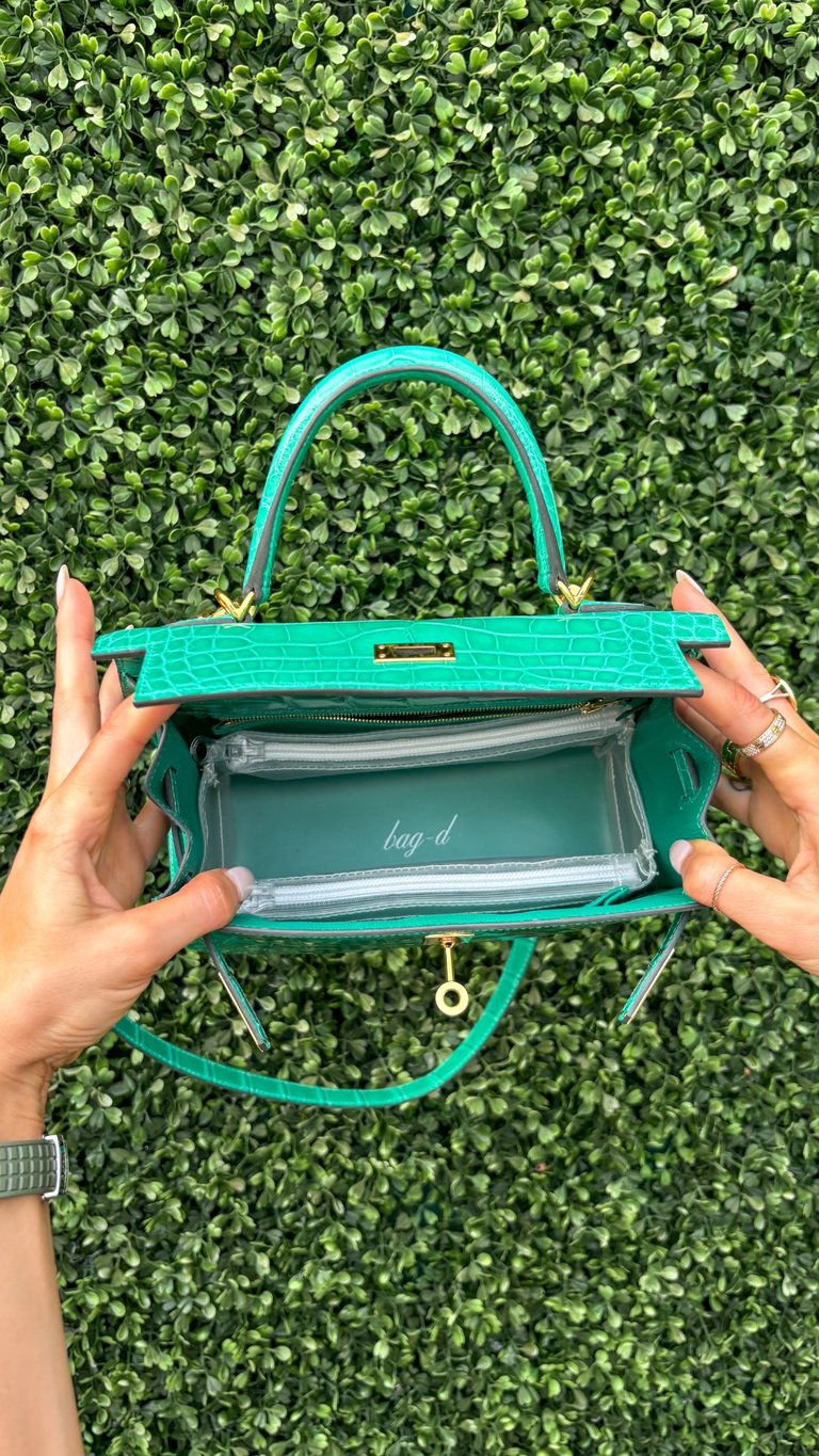 Hands holding an open turquoise crocodile-textured handbag with clear interior against a green leafy background