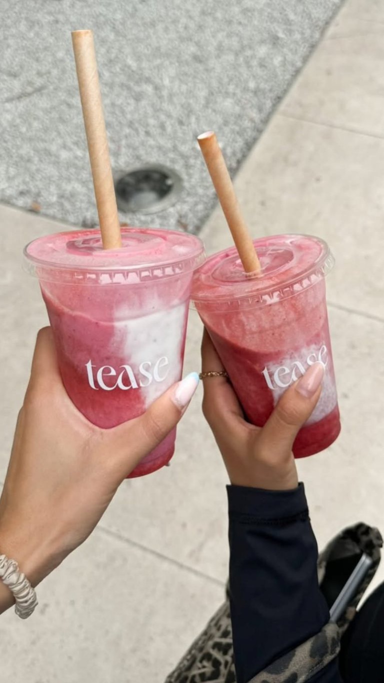 Two hands holding pink Tease coffee drinks with wooden straws outdoors on a gray concrete surface