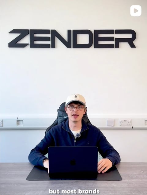 Person wearing a cap and navy hoodie sits at a desk with a laptop in front of the ZENDER logo on a white wall