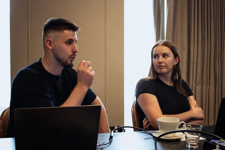 Two people sitting at a table with a laptop and beverages, appearing to be in a discussion or presentation meeting