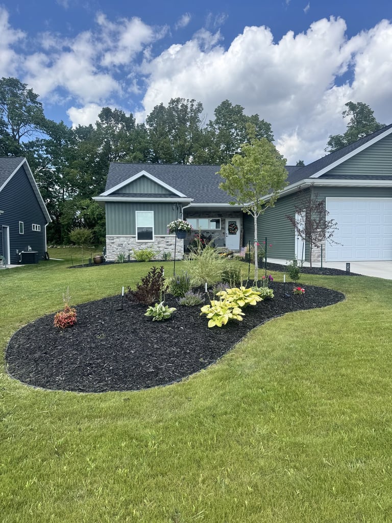 A suburban home with a front yard landscaping bed featuring black mulch, ornamental grasses, and flowering plants under a blue sky with white clouds
