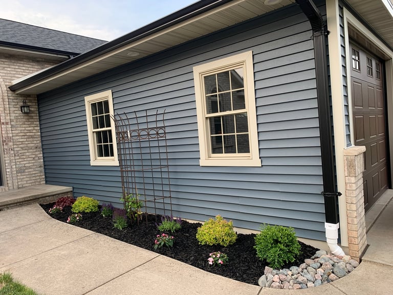 Modern house exterior with blue-gray horizontal siding, cream-colored windows, dark door, and landscaped flower bed with shrubs and decorative stones